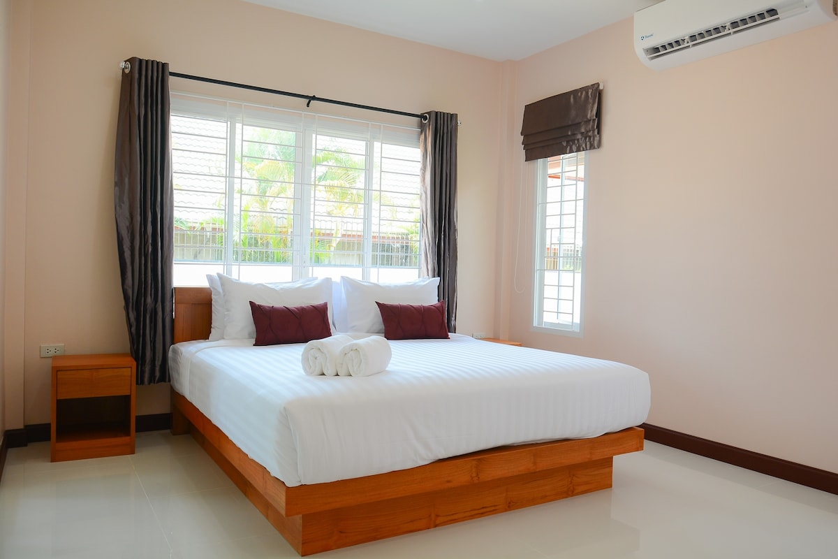 A spacious bedroom is furnished with a large bed featuring white linens and accent pillows. Natural light filters in through large windows, complemented by light curtains. A bedside table stands next to the bed, and an air conditioning unit is mounted on the wall.
