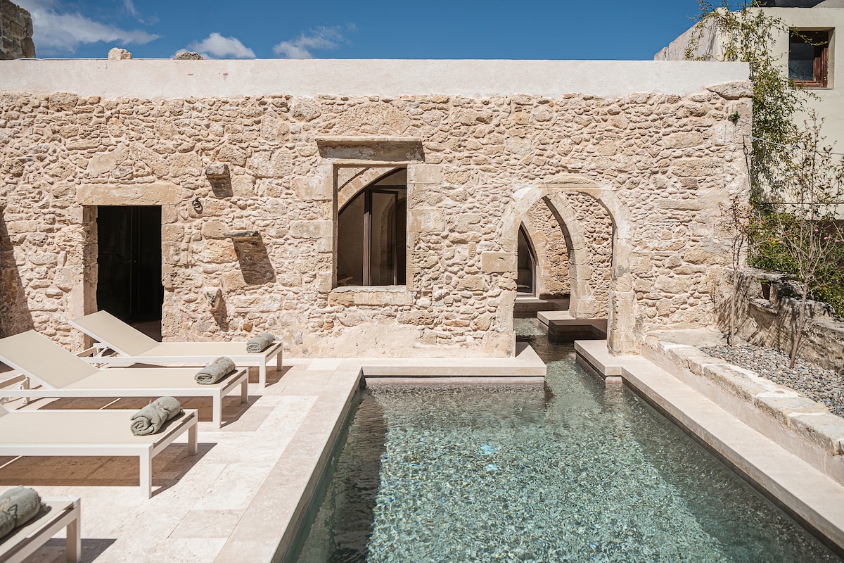A serene outdoor space features a heated semi-covered pool with varying depths, surrounded by natural stone walls. Sunbeds with towels are positioned nearby, inviting relaxation. Arched doorways provide access to the interior, while plants add a touch of nature to the tranquil atmosphere.