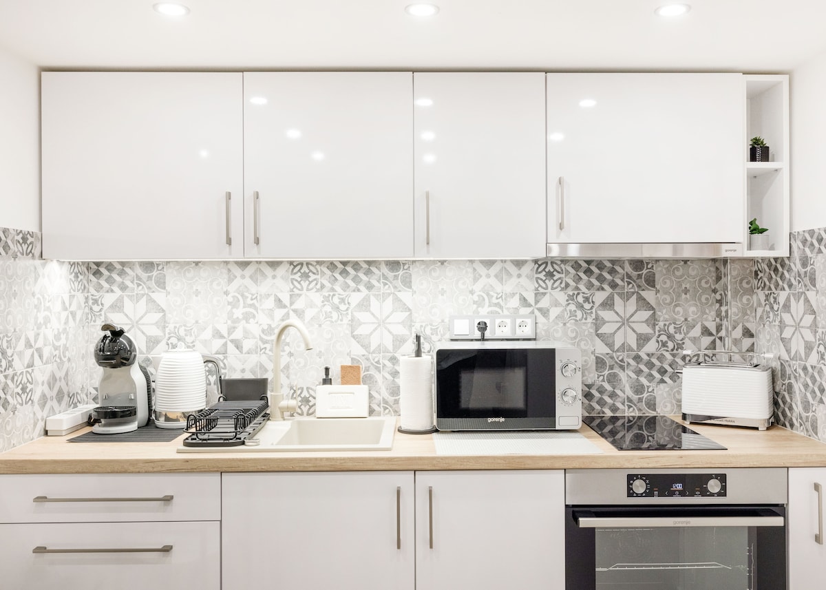 The modern kitchen features a combination of white cabinetry and patterned tiles, providing a stylish backdrop. Essential appliances, including a microwave, kettle, and oven, are organized neatly on the countertop, which is finished in light wood. Ample lighting illuminates the space.