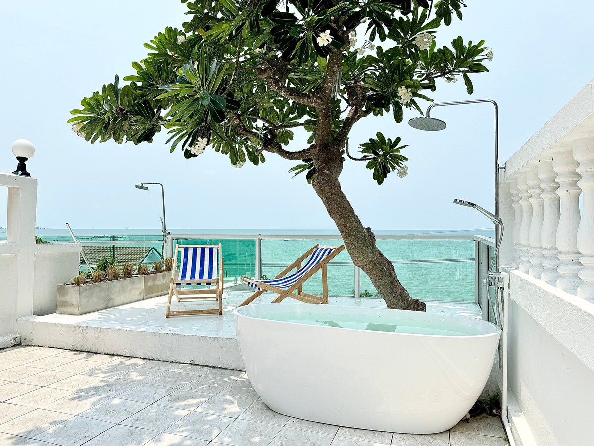 An outdoor terrace features a freestanding white bathtub surrounded by green foliage. A tree provides natural shade, while a pair of lounge chairs with striped fabric offer relaxation options. The panoramic ocean view enhances the serene ambiance of the space.