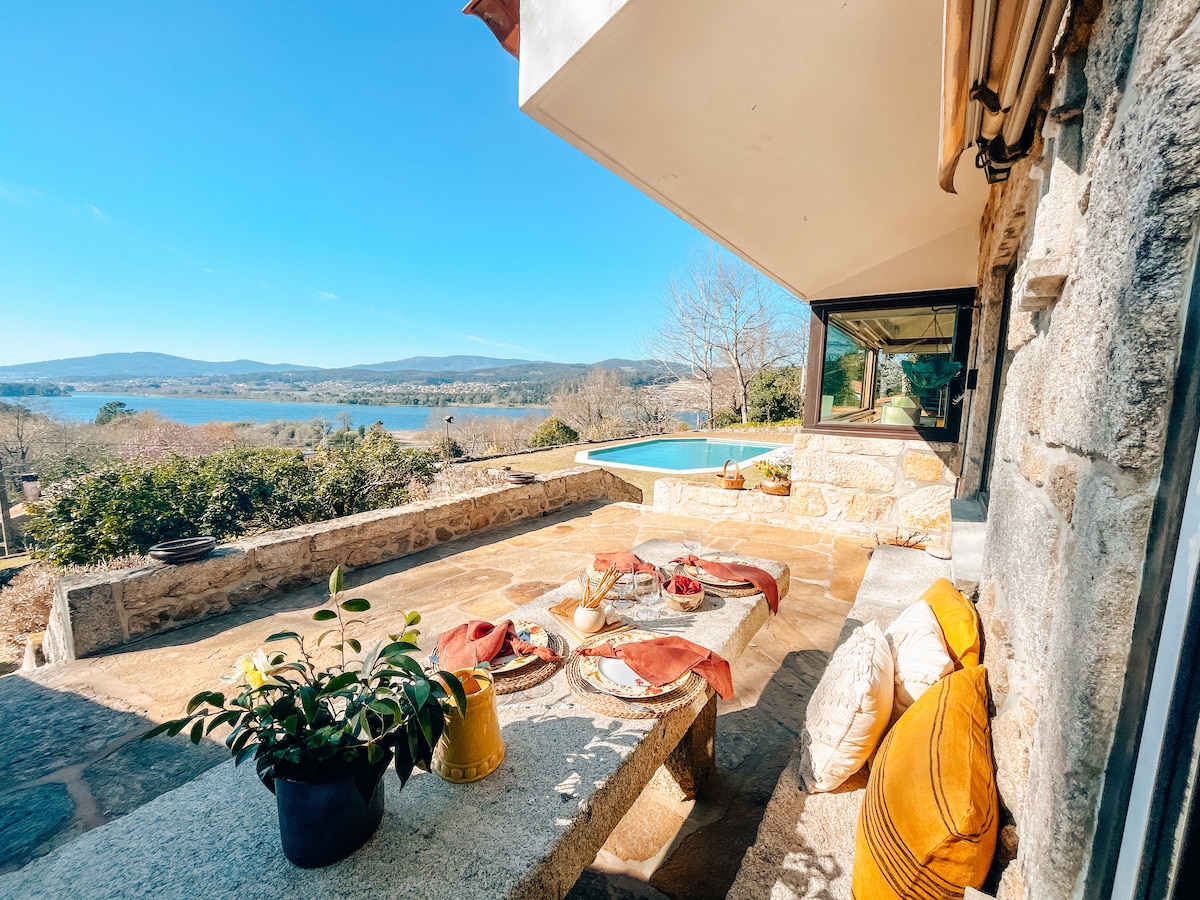 An outdoor terrace merges with the scenic landscape, featuring a stone table adorned with colorful textiles and cushions. A swimming pool can be seen in the background, with expansive views of the river and hills under a clear blue sky.