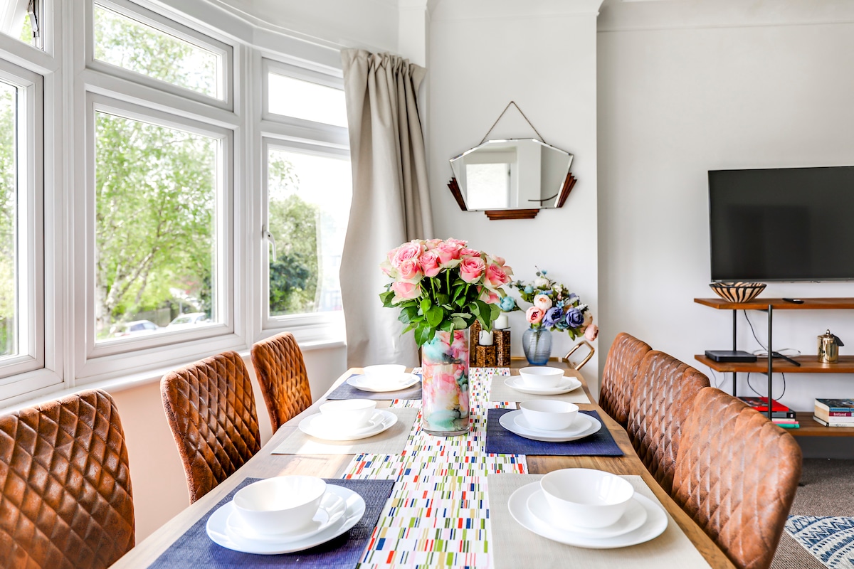 A dining area features a long table set for six, with white plates resting on blue placemats. A colorful runner decorates the table, adorned with a vase of pink flowers and a mirror hanging on the wall. Soft light enters through large windows, enhancing the space.