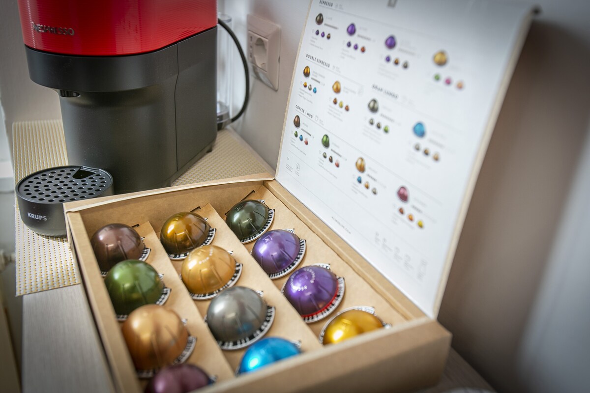 An espresso machine in bright red sits on a countertop beside an assortment of coffee capsules. The capsules are housed in a neatly arranged box, showcasing various colors, indicating a selection of flavors. A detailed chart of the capsule varieties is positioned nearby.