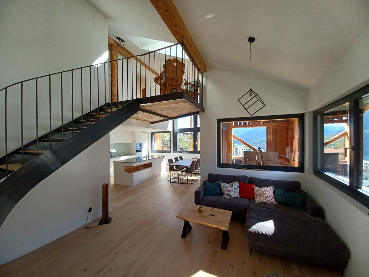 A spacious living area features large windows allowing natural light to fill the room. A comfortable sectional sofa is arranged around a wooden coffee table. A modern staircase leads to an upper level with dining seating visible, and an open kitchen is situated in the background.