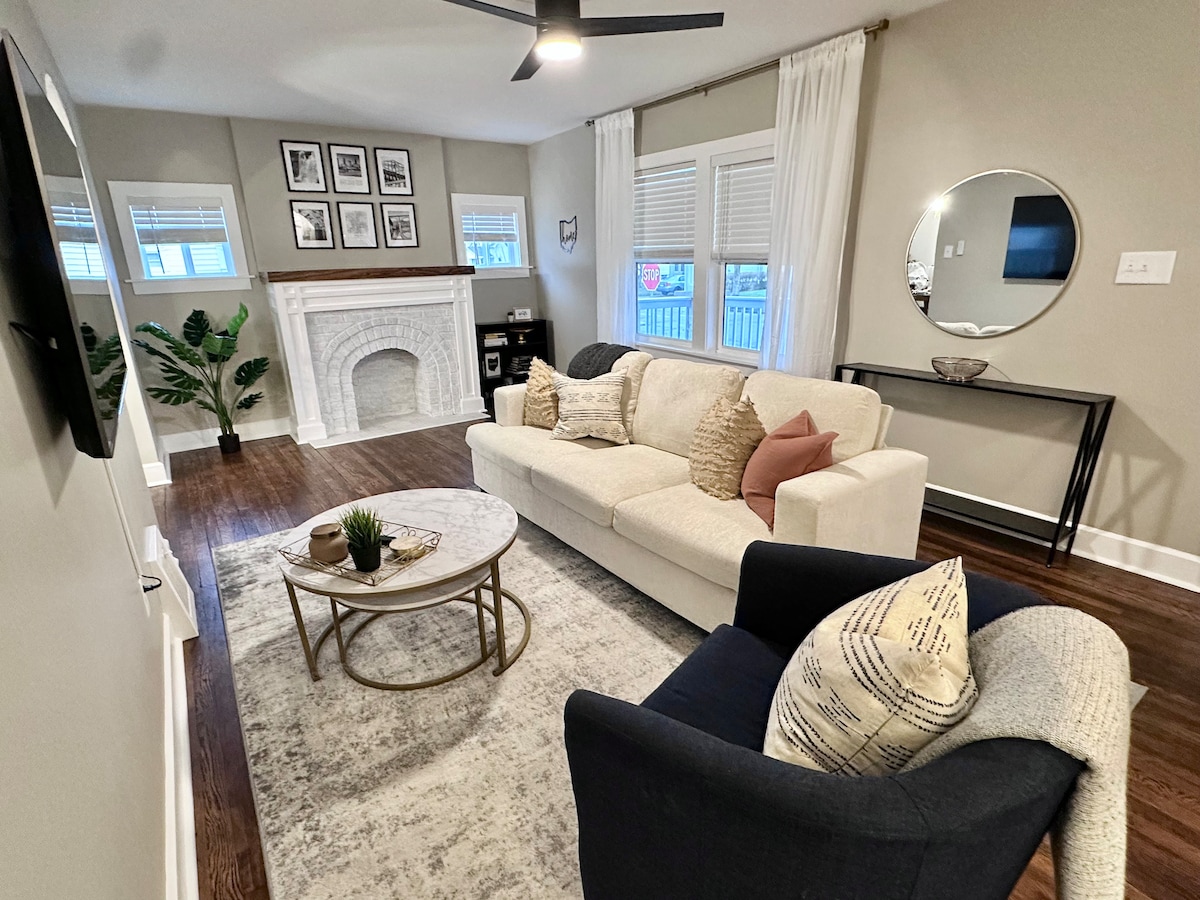 A cozy living area features a light-colored sofa adorned with decorative pillows, arranged around a circular coffee table. A stylish armchair with geometric patterns complements the space. Natural light flows through the window treatments, and a decorative fireplace adds character to the room.
