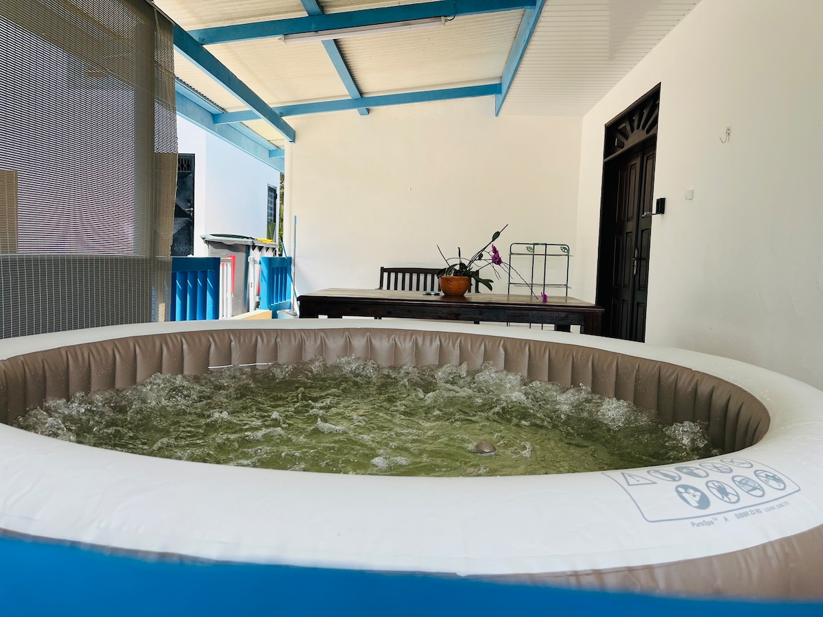 A spa is positioned on the terrace, showcasing a circular design with bubbles visible in the water. Seating is available nearby at a wooden table, complemented by an indoor plant. The walls are a soft-toned color, and clear skies are visible beyond the structure.