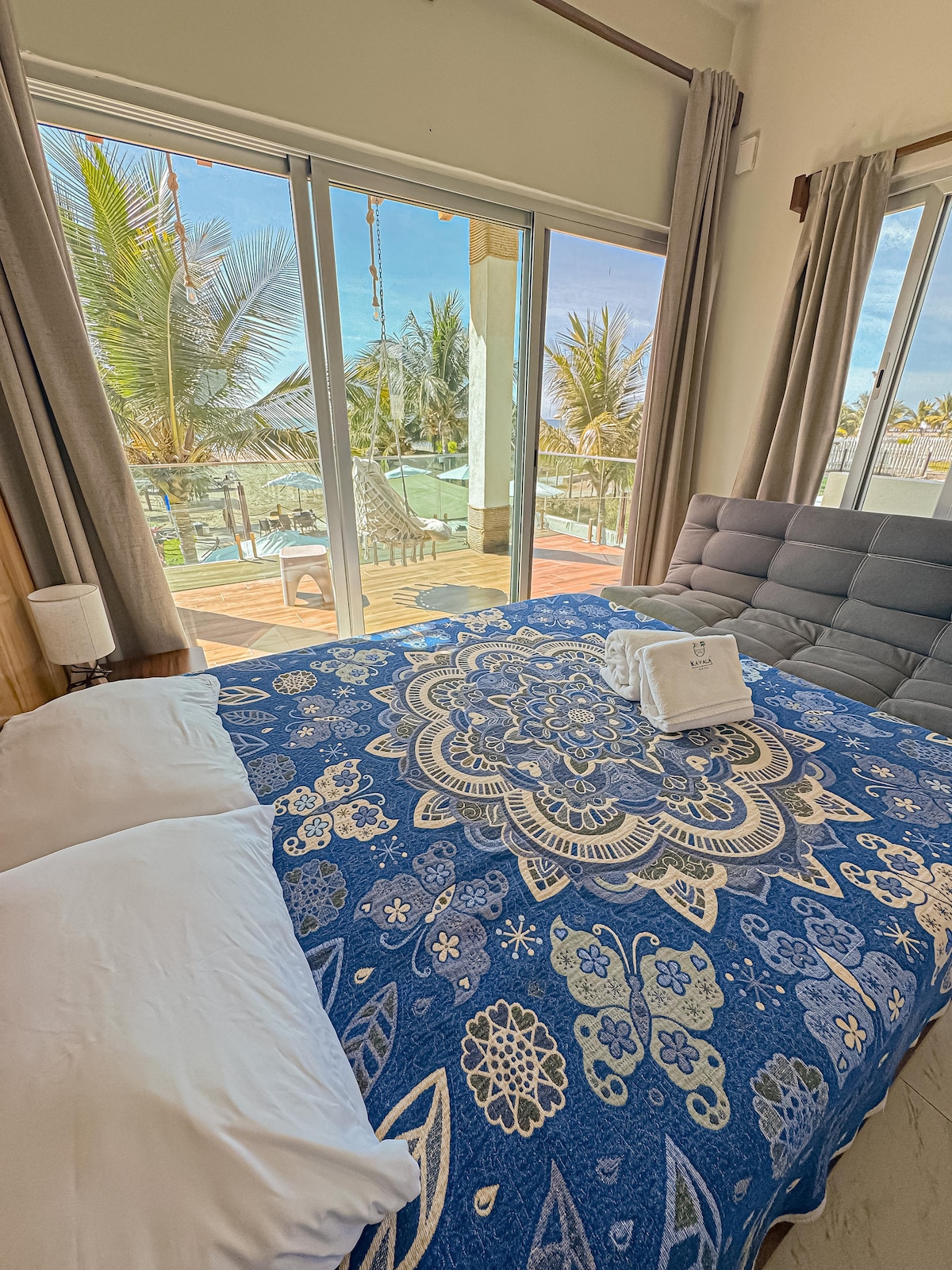 A spacious bedroom features a large bed covered with a blue patterned blanket. Floor-to-ceiling glass doors provide access to a private outdoor area surrounded by palm trees, allowing natural light to fill the room.