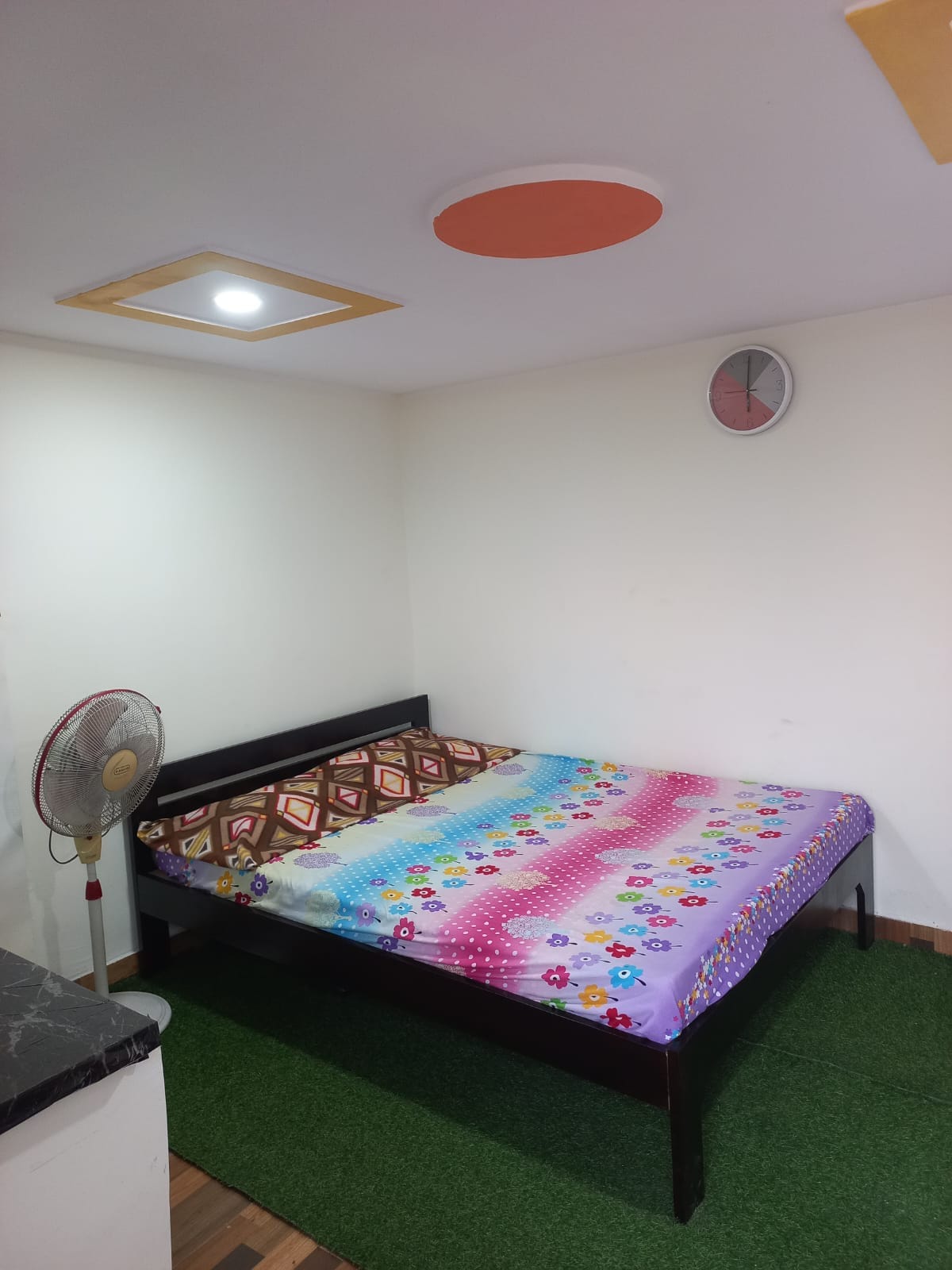A cozy sleeping area features a spacious bed adorned with a colorful patterned blanket. A small fan is positioned nearby, and a wall clock is visible above. Soft green flooring adds a touch of warmth to the minimalist design.