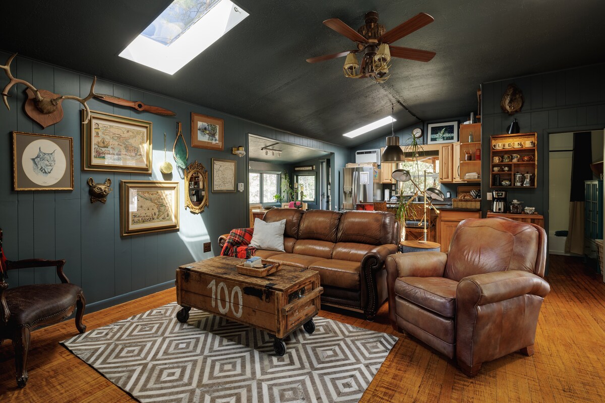 The living room features a spacious layout with leather sofas and a decorative coffee table. A large skylight allows natural light to illuminate the space, complemented by rustic decor on the walls. A cozy area rug adds warmth to the wooden floor.
