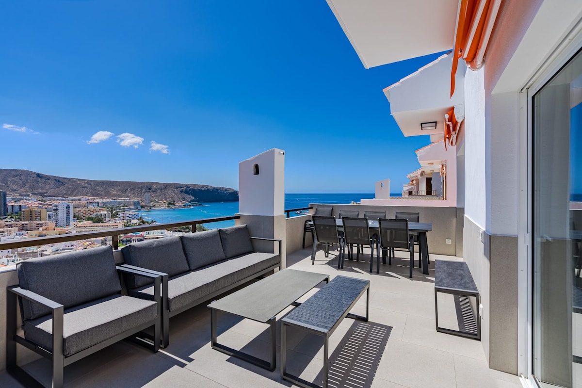 A spacious terrace offers panoramic ocean and city views, furnished with a large seating area, dining table, and shaded areas. The modern design features neutral tones, while the vibrant blue sky complements the coastal surroundings.