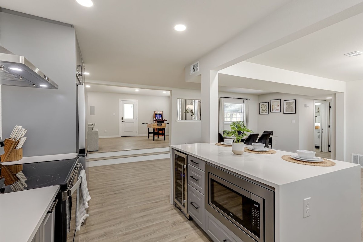 A modern kitchen area is shown, featuring a large island with a microwave and built-in storage. Natural light fills the space through nearby windows, which reflect a minimalistic design. In the background, an open living area is visible, showcasing comfortable seating and a neutral color palette.
