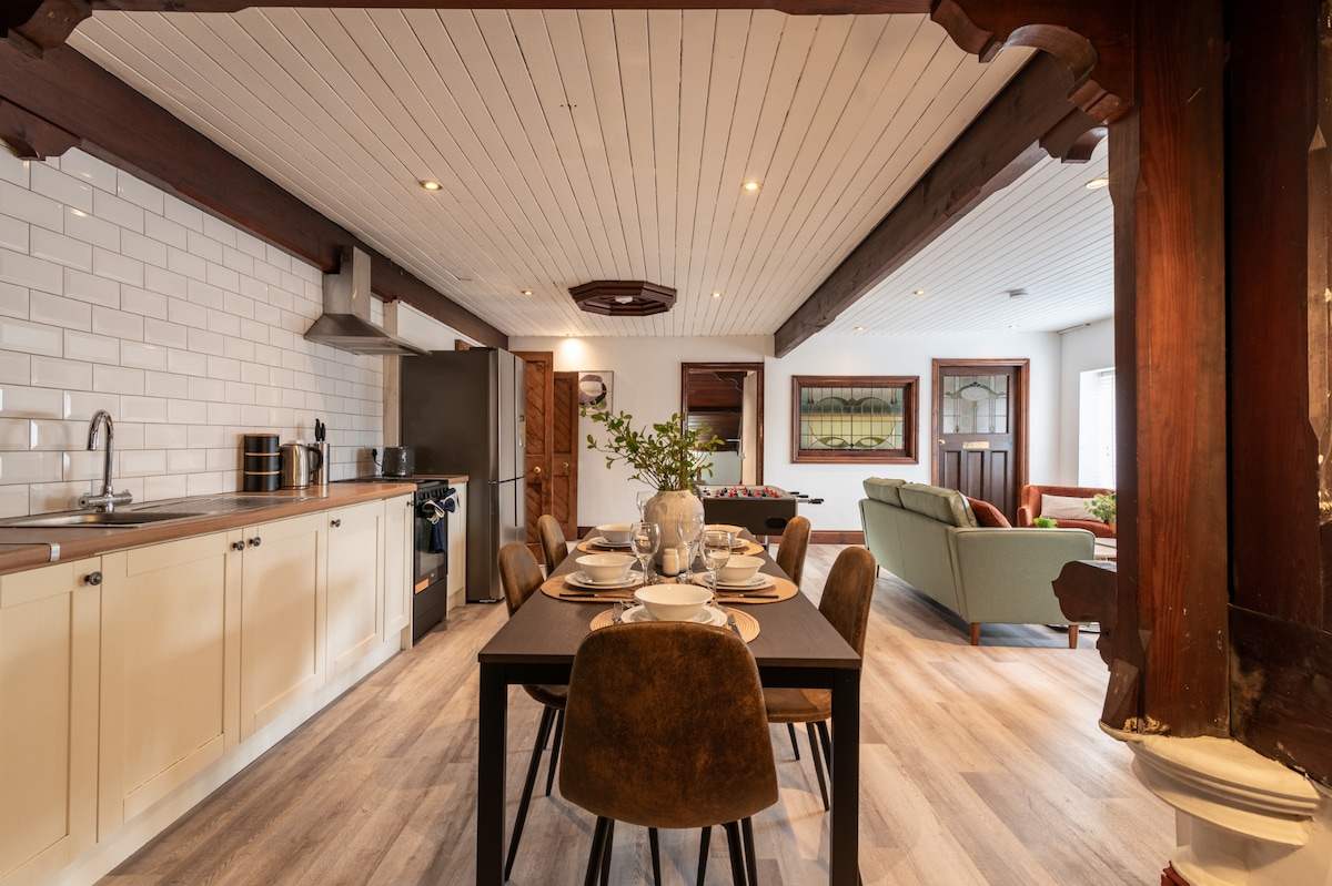 The dining area features a dark wood table set for six, surrounded by modern brown chairs. A well-equipped kitchen is visible in the background, with sleek cabinetry and stainless steel appliances. Natural light fills the space through windows, enhancing the open layout.
