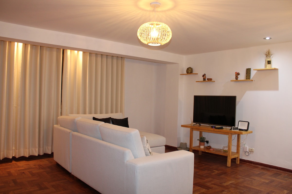 A bright living area features a light-colored sofa facing a wooden entertainment unit with a television. Shelves on the wall display decorative items. Sunlight filters through sheer curtains, enhancing the spacious feel of the room, which is complemented by warm wooden flooring.