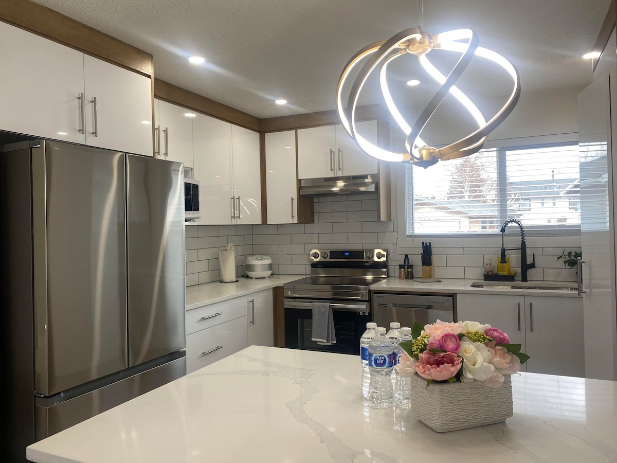 The modern kitchen showcases stainless steel appliances, including a refrigerator and oven, situated against a sleek backsplash. A centrally located island features a stylish light fixture above, and fresh flowers in a decorative bowl add a touch of color. Natural light filters in through the window.