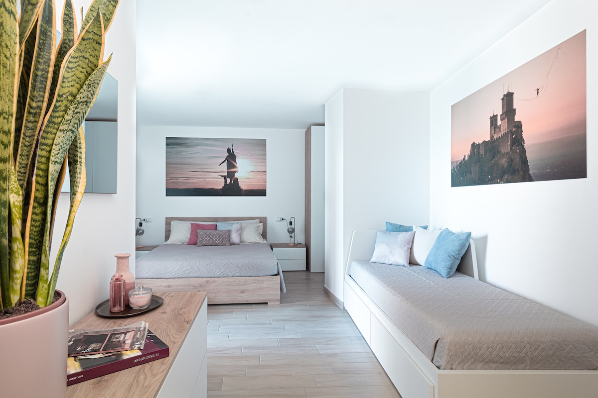 A spacious bedroom is presented featuring two beds, one being convertible, and artwork depicting a mountain landscape. Natural light enters through a nearby window, enhancing the modern decor. A small bedside table and decorative elements, including a plant, add warmth to the space.