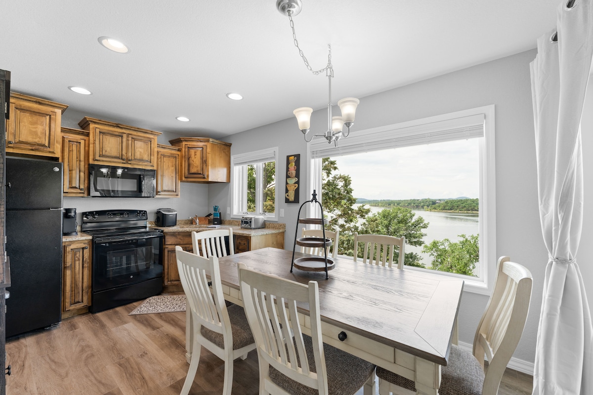 A bright dining area is featured, furnished with a wooden table and white chairs. Natural light floods in through large windows, providing a view of the lake and surrounding greenery. The kitchen area, equipped with modern appliances and wooden cabinetry, is seamlessly integrated into the space.