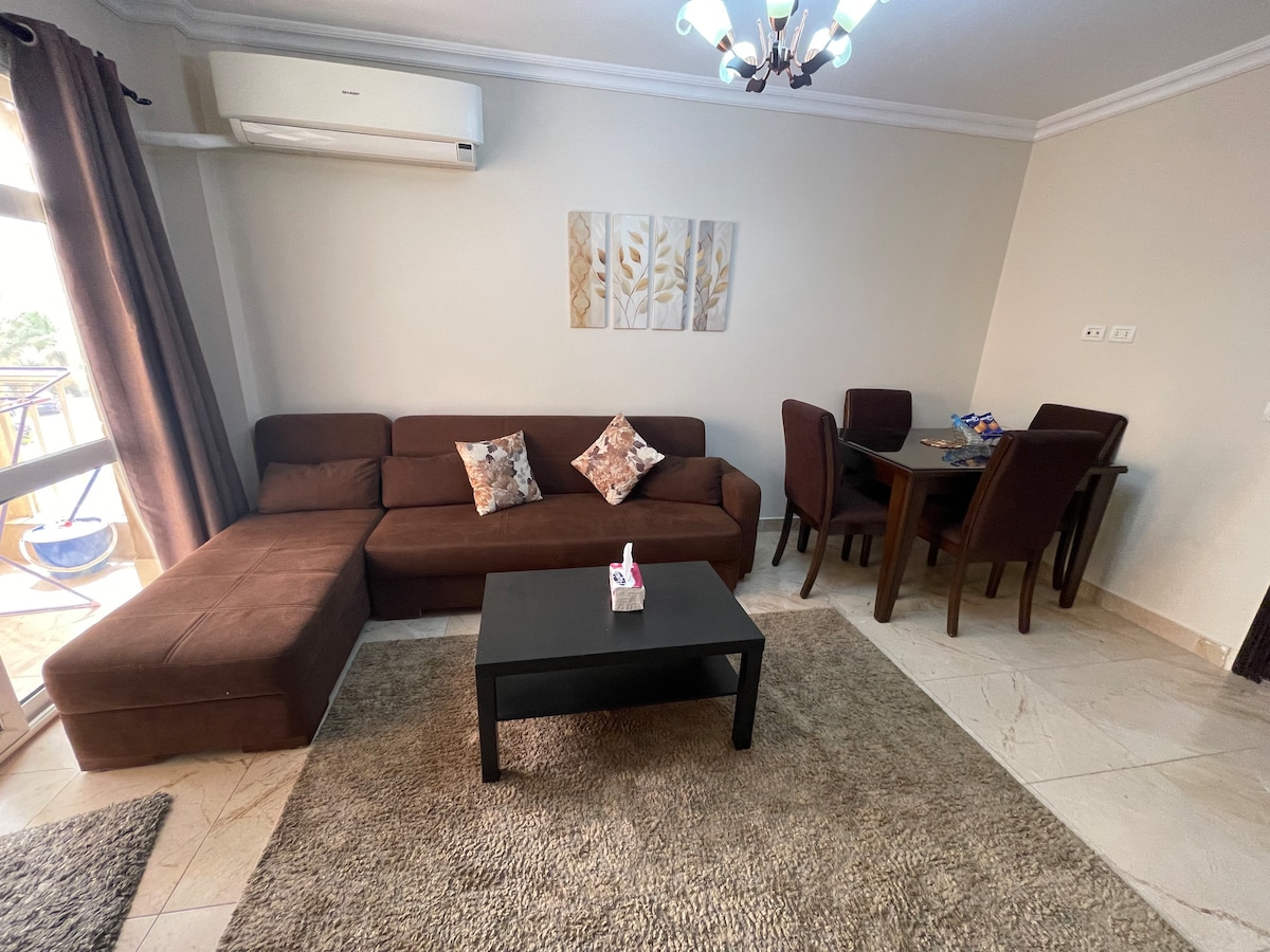 A cozy living space is showcased, featuring a dark brown sectional sofa adorned with decorative pillows. A small coffee table is positioned in front, with a soft area rug underneath. A dining table with four chairs is visible in the background, beside a window dressed with light curtains.