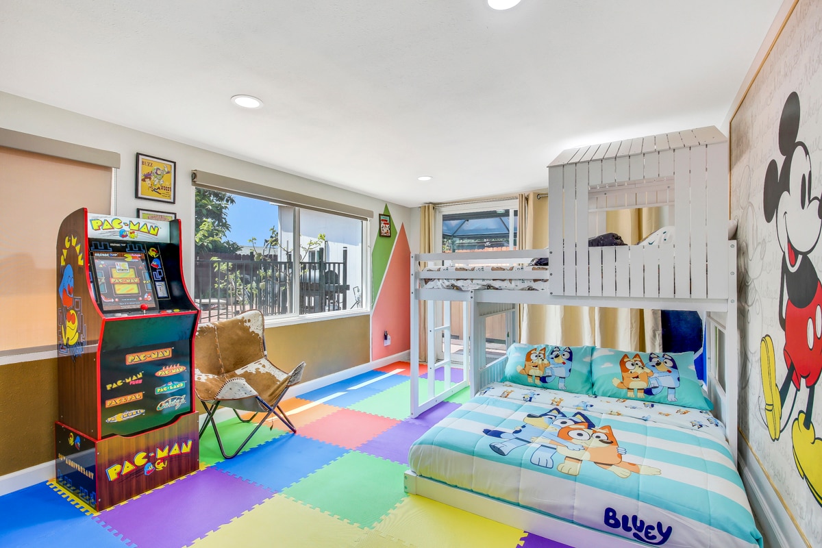 The kid's room features a bunk bed with a lower sleeping area and colorful Bluey-themed bedding. A classic arcade game machine adds a fun element, while the brightly colored foam floor tiles create a safe play environment. Natural light enters through large windows.