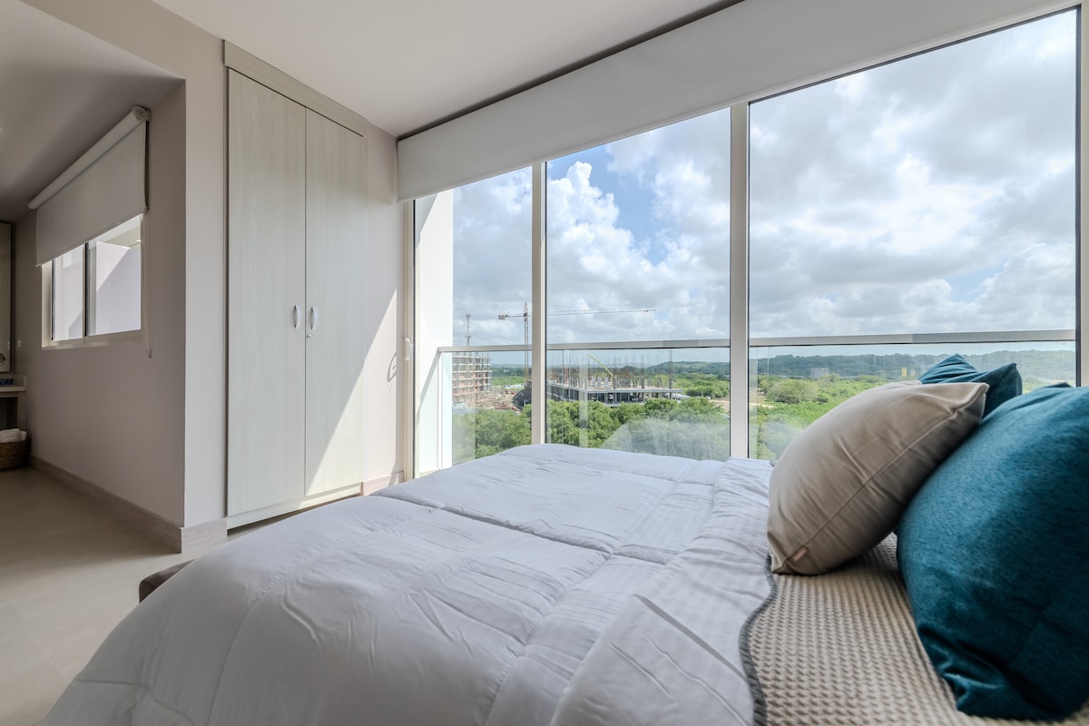 A spacious sleeping area features a queen bed, adorned with soft pillows. Large glass windows offer a view of lush greenery and a serene sky, while ample natural light brightens the room. A modern wardrobe is positioned beside the bed, enhancing the room's functionality.