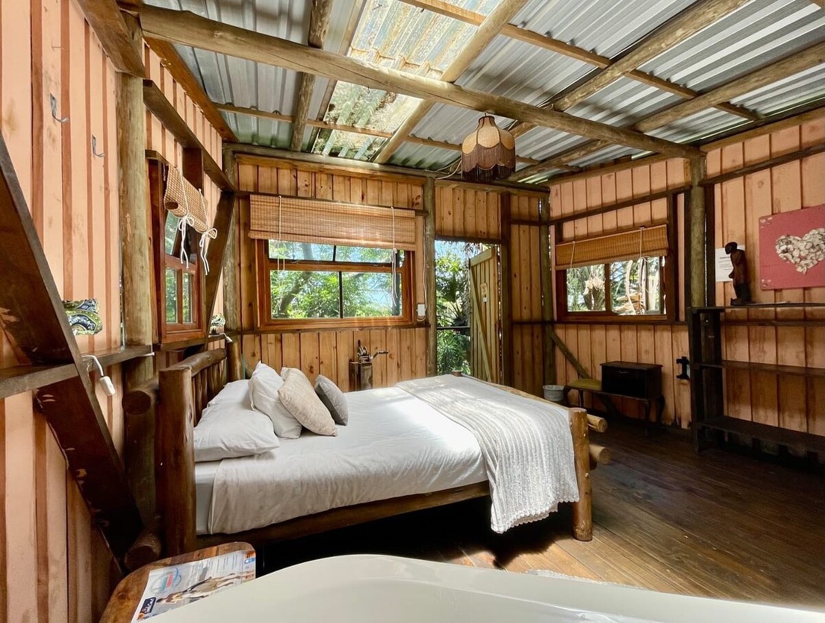 A rustic bedroom features a spacious bed adorned with neutral-toned pillows and a textured blanket. Large windows invite natural light and showcase the surrounding greenery. The wooden walls and ceiling enhance the room's natural aesthetic, complemented by warm-toned light fixtures.