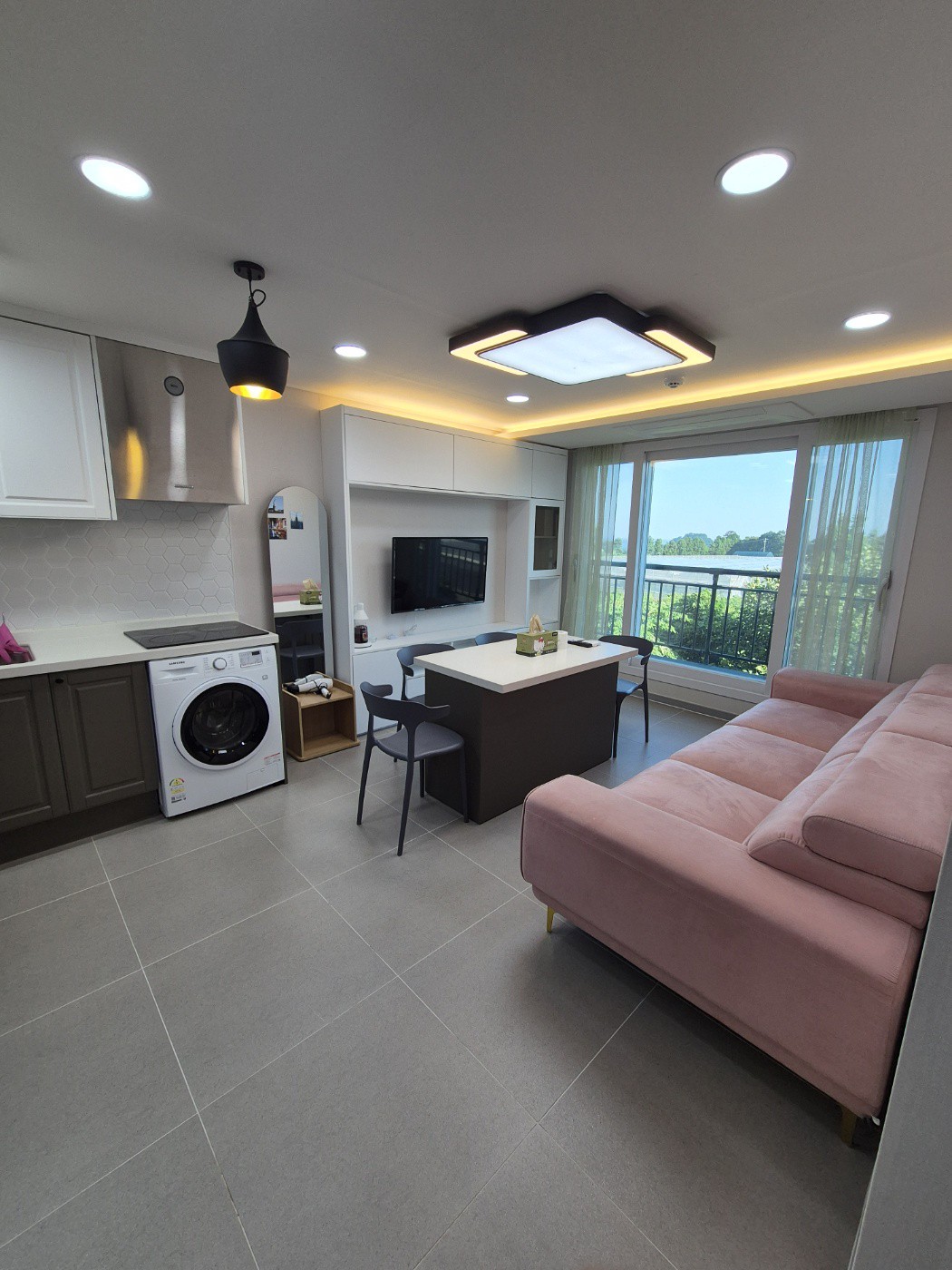 The living space features a modern kitchenette with a washing machine, an entertainment area with a wall-mounted television, and a cozy dining setup. A soft pink sofa creates a comfortable seating arrangement, while large windows allow natural light and offer views of the surrounding landscape.
