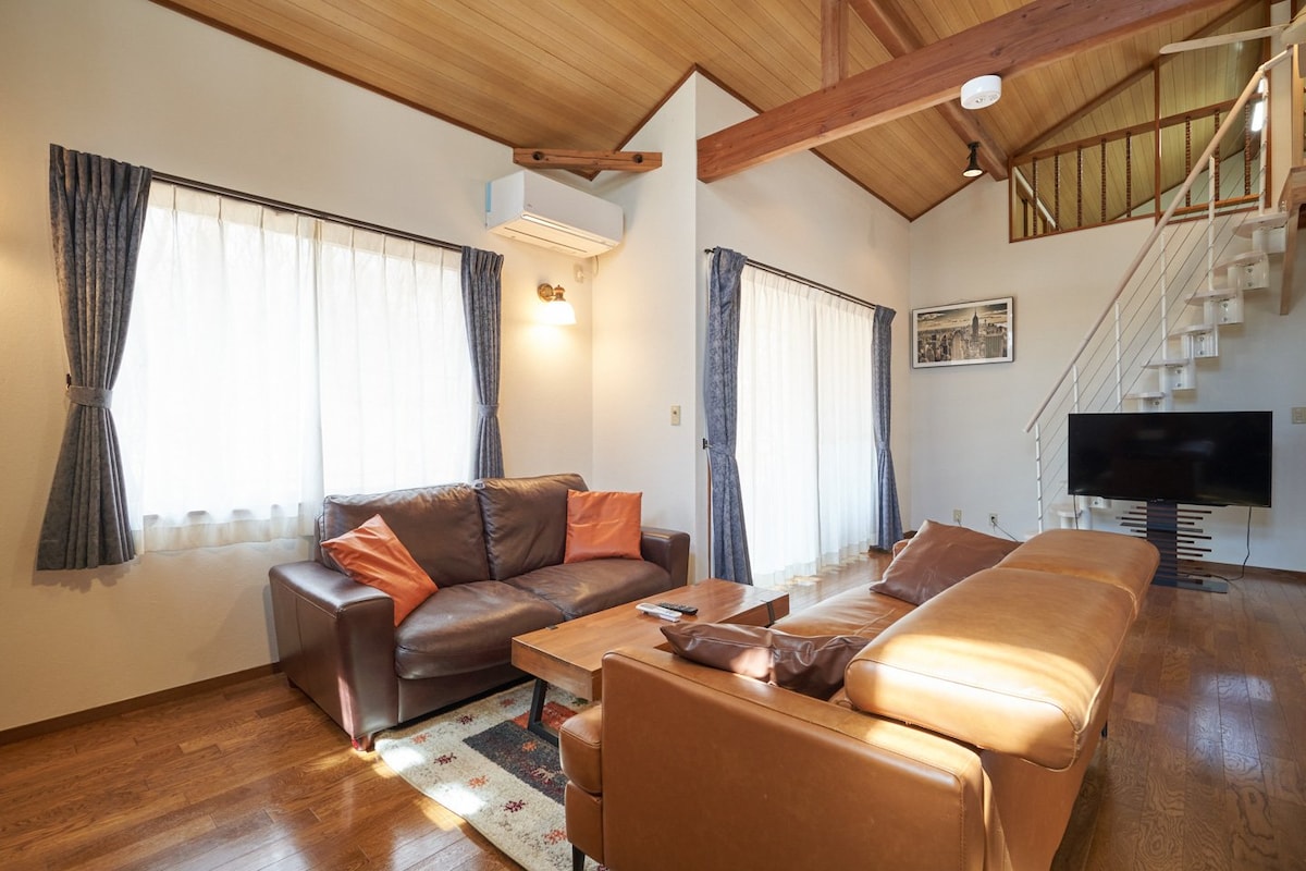 A living area is featured with large windows allowing natural light to fill the space. Comfortable leather couches, one adorned with orange pillows, surround a central wooden table. A wall-mounted television is positioned nearby, creating an inviting space for relaxation and gathering.