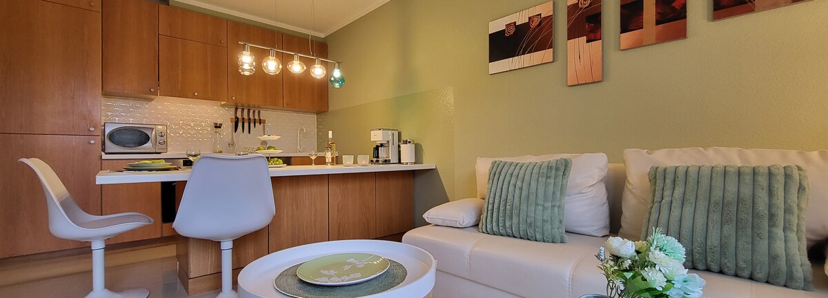 A modern kitchen area is visible, featuring wooden cabinetry and a breakfast bar. Soft green walls complement white furniture, which includes a sofa adorned with decorative pillows. A small table is set with a plate, while stylish light fixtures hang above the space, enhancing the cozy atmosphere.