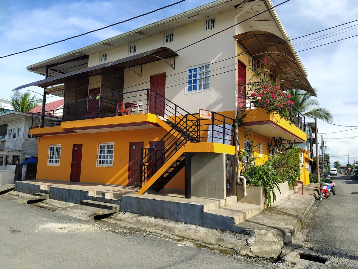 The exterior of a multilevel building features bright orange accents and a staircase leading to upper units. Balconies are adorned with small tables and chairs, enhancing outdoor relaxation. The building is situated on a quiet street, with palm trees and utility lines framing the scene.
