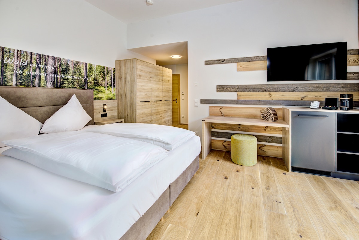 A modern bedroom features a spacious bed with white linens and decorative pillows. Natural wood accents complement the contemporary design. A flat-screen television is mounted on the wall above an elegant console, which includes a coffee station. Soft lighting enhances the welcoming atmosphere of the room.
