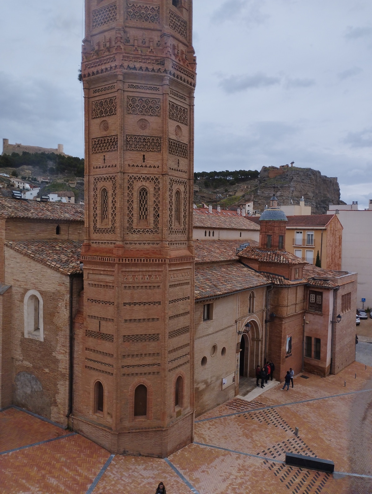 A tall brick tower features intricate patterns and textures, standing alongside a historic building with arched windows. The surrounding square is paved, inviting visitors to explore. A rocky hill rises in the background, contrasting with the structured architectural designs.