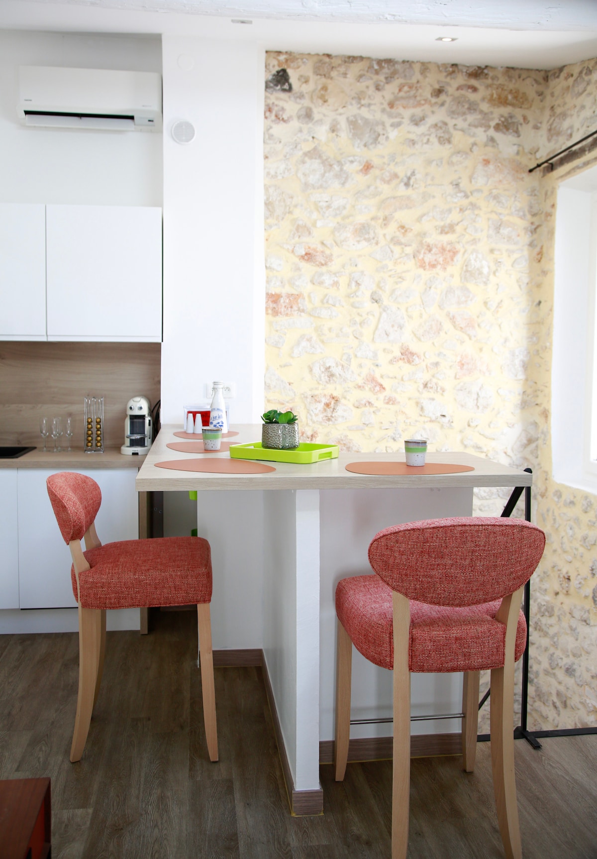 A kitchen area is presented with a minimalist design, featuring a stone wall and modern cabinetry. Two bar stools with red upholstery are positioned at a small white counter, accommodating a simple table setting with a potted plant and drink. Natural light illuminates the space.
