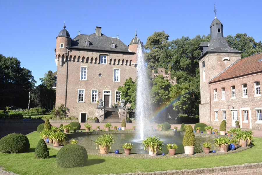 A historic castle is set amidst lush greenery, featuring a central fountain surrounded by neatly trimmed hedges and potted plants. The castle's stone façade displays towers and arched windows, while the clear blue sky complements the tranquil environment.
