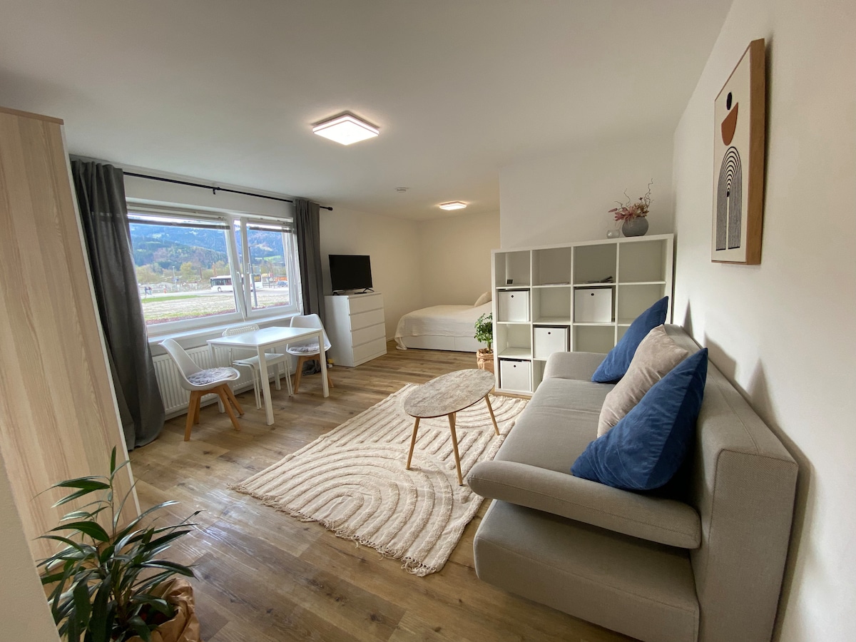 A modern living space is presented, featuring a comfortable seating area with a light gray sofa and blue accent pillows. A compact dining area with two tables and chairs is visible. Natural light floods in from a large window, illuminating the wooden floors.