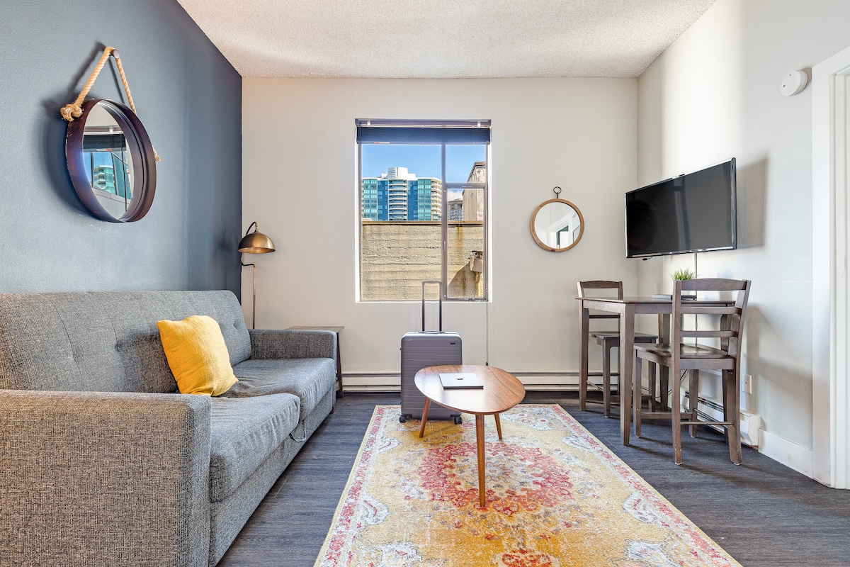 The living area is furnished with a gray sofa, a round coffee table, and a dining space featuring two high-backed chairs. Natural light enters through a large window, illuminating the room's neutral tones. A small suitcase is placed beside the sofa, with decorative mirrors on the walls.