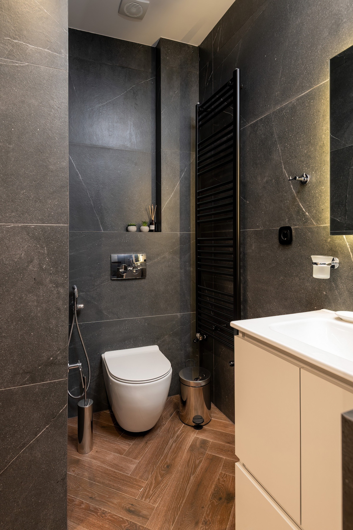 A modern bathroom is displayed with sleek black stone walls and warm wooden flooring. A compact toilet is positioned against one wall, complemented by a stylish towel radiator. A minimalist sink area features a simple design, enhancing the overall clean aesthetic.