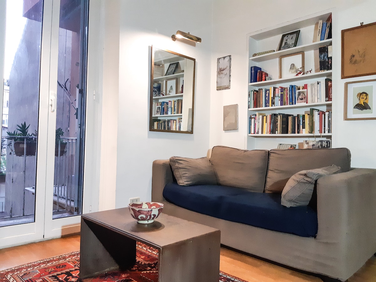 A living area features a comfortable sofa with dark blue accents, positioned across from a wooden coffee table. A bookshelf filled with books is visible along one wall, while a mirror reflects light from a nearby window that opens to a small balcony.