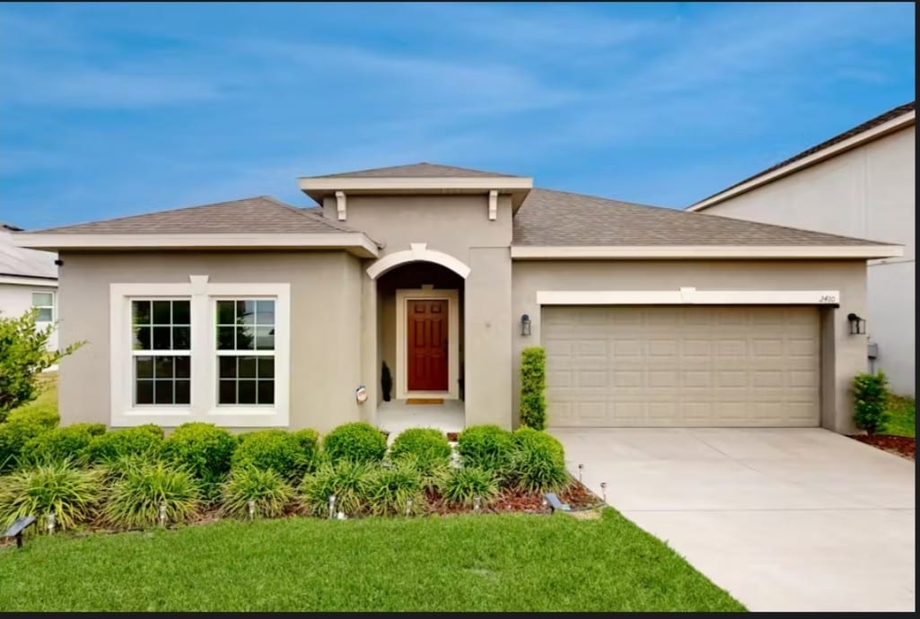 A single-story home is featured with a neatly landscaped front yard, showcasing a variety of green shrubs and grass. The entrance features a covered porch and a wooden front door, while two large windows provide a view into the living space. A driveway leads to a two-car garage.