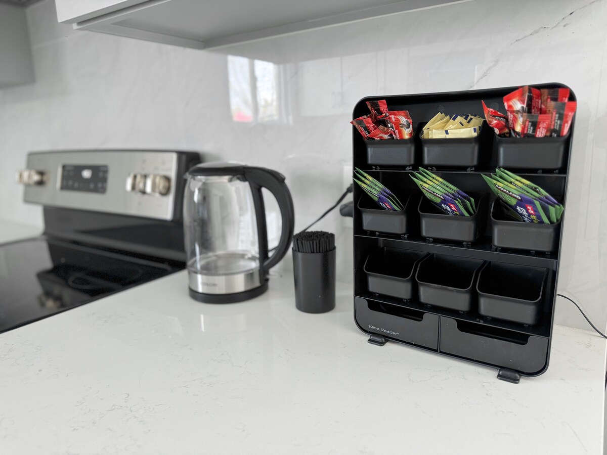 A modern kitchen counter features a stylish tea and coffee station, with various packets of tea and coffee neatly organized in a black holder. A kettle is positioned nearby, alongside a collection of disposable cups, set against a sleek marble backdrop.