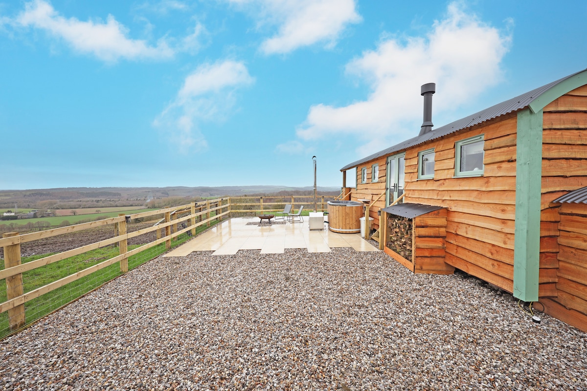 An outdoor patio area is presented with a mix of gravel and smooth stone flooring. A woodfired hot tub is positioned next to the shepherd's hut, and comfortable seating is available for relaxation. Expansive views of the surrounding landscape are visible in the distance.