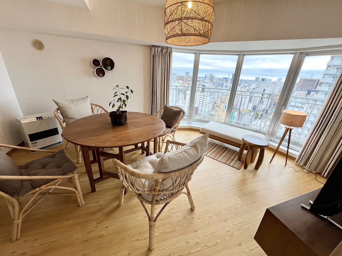 A bright dining area features a round wooden table surrounded by four chairs, including two with woven backs. Large windows offer panoramic views of the city, allowing natural light to fill the space. A small plant sits atop the table, enhancing the inviting atmosphere.