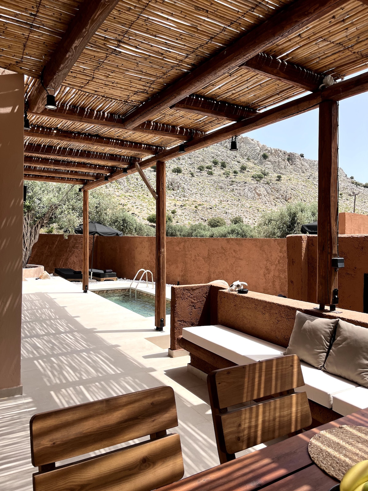 A shaded terrace area is presented, featuring a wooden dining table and a cozy seating arrangement. The private pool is visible in the background, surrounded by natural stone walls and olive trees, with sunlight casting intricate shadows across the space.