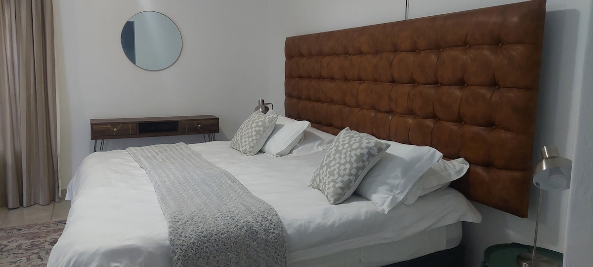 The bedroom features a large, tufted leather headboard that complements the spacious bed topped with white linens and decorative pillows. A circular mirror hangs above a modern wooden dresser, while soft curtains frame the window, allowing natural light to brighten the room.