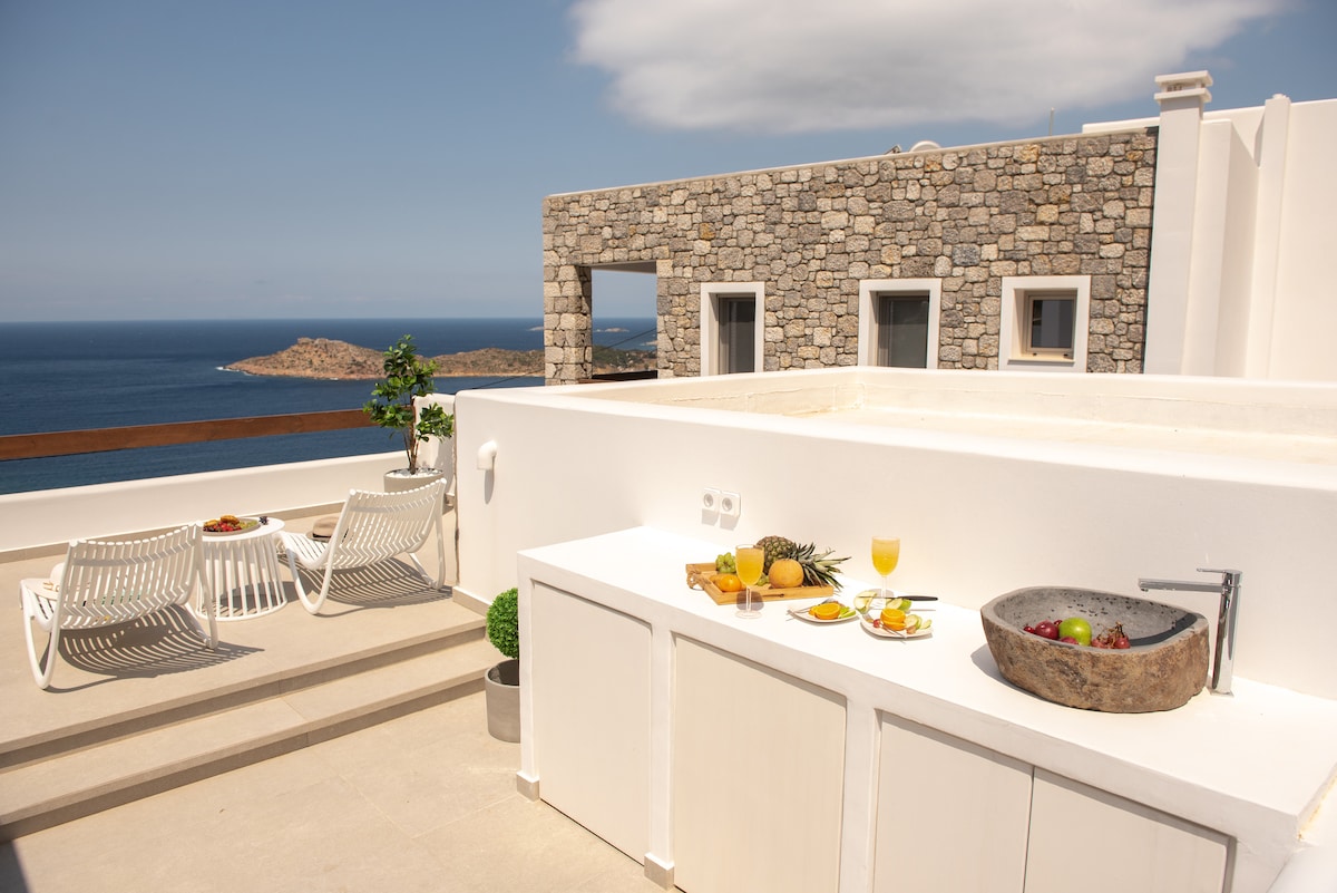 An outdoor terrace is presented, featuring a kitchenette with a stone sink and a wooden serving tray adorned with colorful fruits. White lounge chairs are positioned nearby, offering a view of the sea and distant island, under a clear sky.