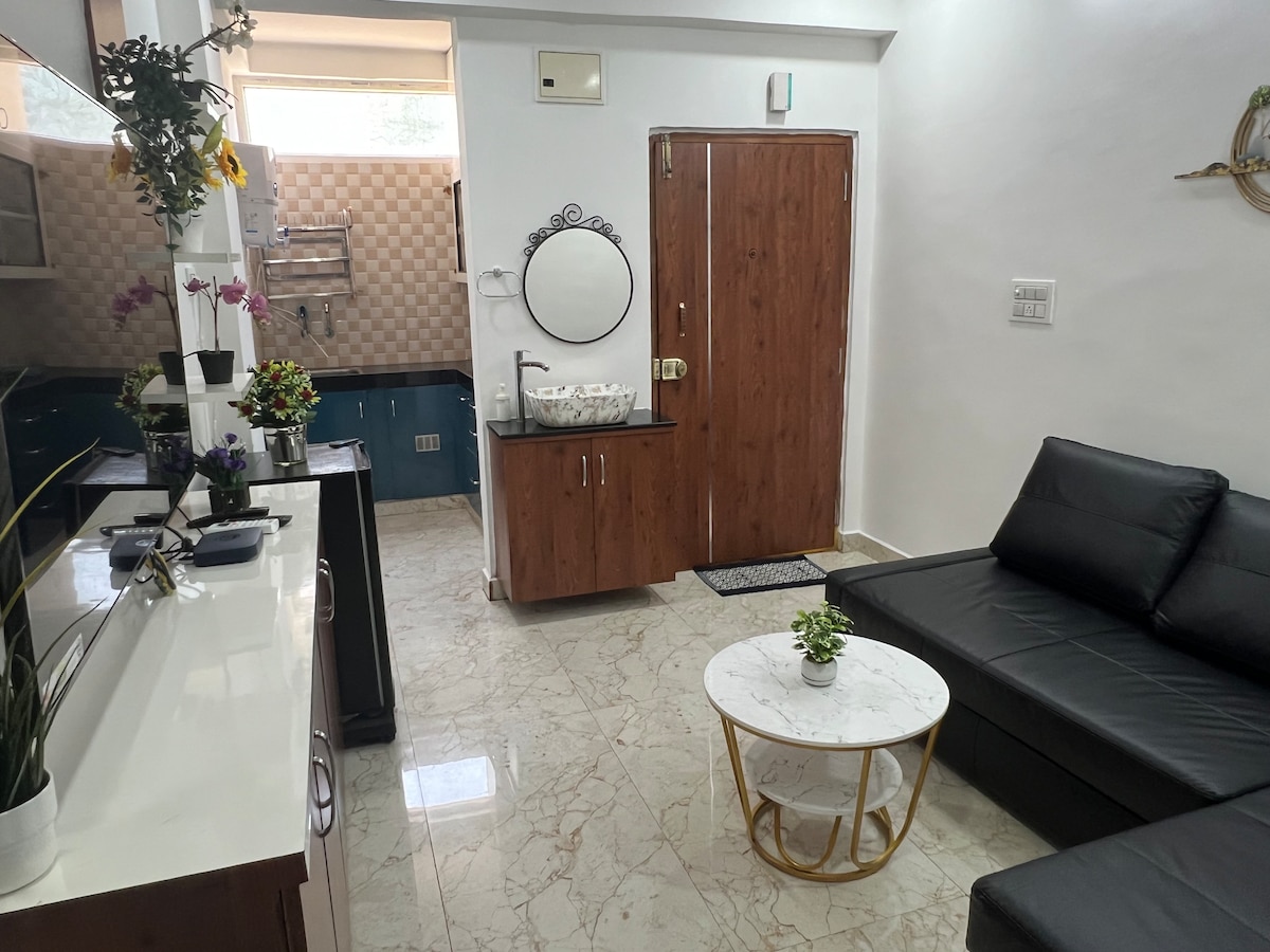 A modern living space is shown, featuring a sleek black sofa paired with a round marble coffee table. A small plant adorns the table. The kitchen is visible in the background, along with a mirror and decorative items on the wall.