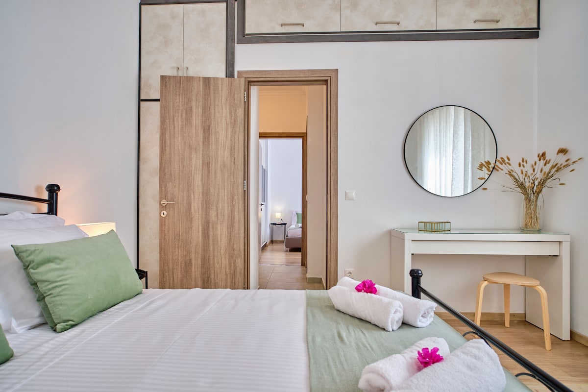 A bright sleeping area features a spacious bed adorned with green pillows and neatly rolled towels. A round mirror hangs above a simple desk, reflecting soft natural light. The open door leads to another room, enhancing the sense of space.