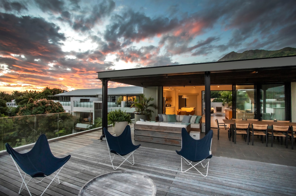 A spacious terrace is visible, featuring modern outdoor seating arrangements. The expansive view captures a vibrant sunset with a mix of orange and purple hues. Ample indoor living space can be seen through large glass doors, inviting natural light to flow in.