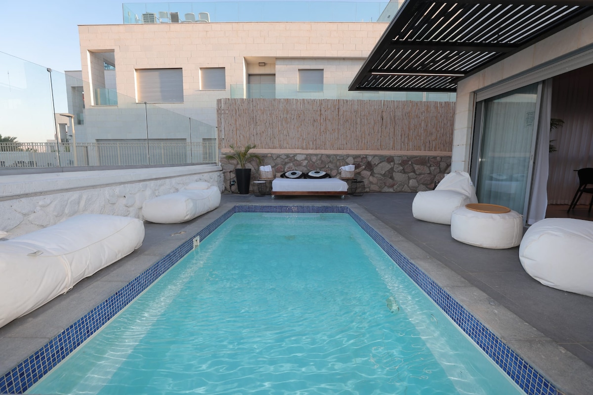 A private outdoor area is captured, featuring a serene swimming pool surrounded by soft white seating. A sunbed is positioned nearby, offering a place to relax under the open sky. Natural stone and wood elements enhance the tranquil atmosphere, with a modern architectural design in the background.