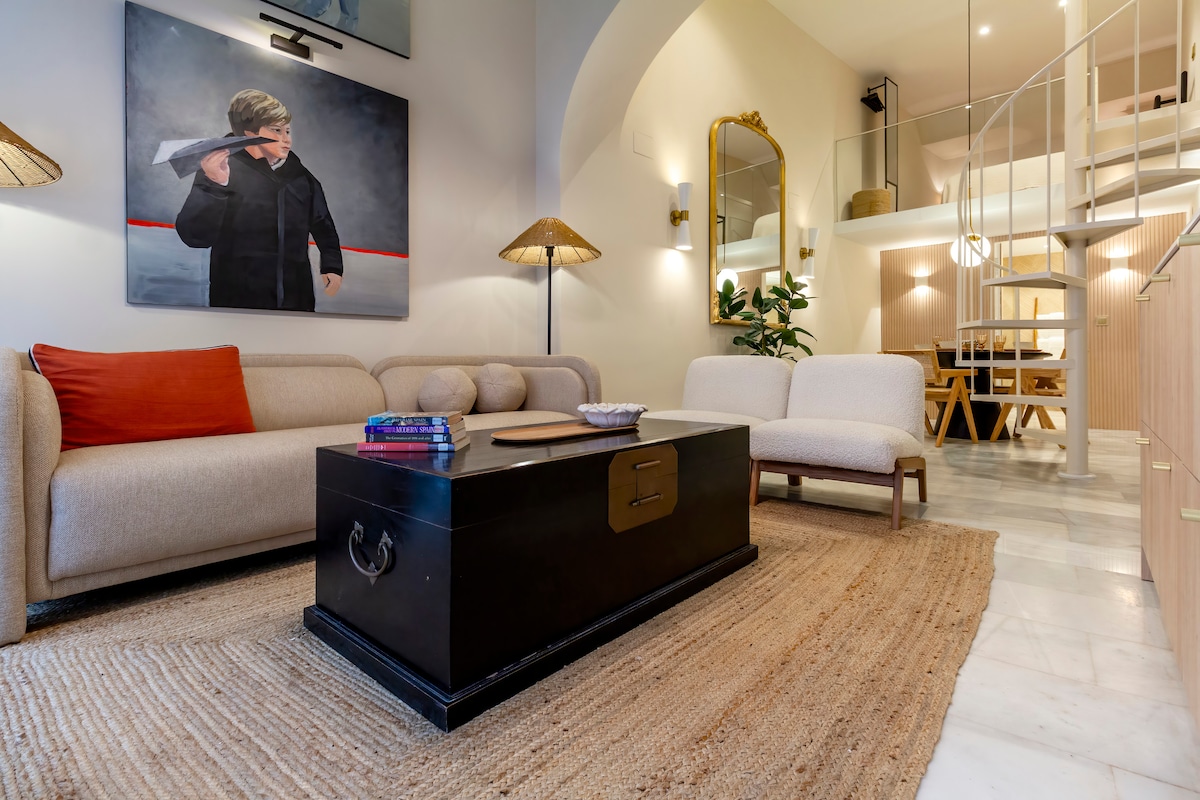 The open plan living area features a large, soft sofa accompanied by two armchairs. A coffee table is centrally placed on a textured rug. Natural light enters through the high ceiling, complementing the contemporary art piece on the wall and the decorative elements.