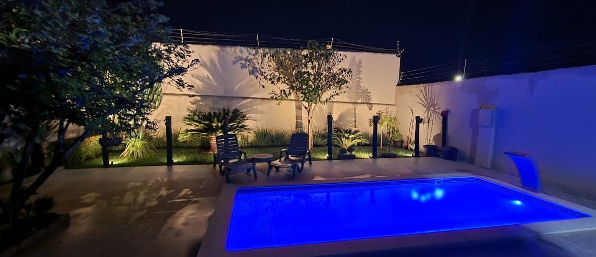 A tranquil outdoor space is presented at night, featuring a softly illuminated pool with a calming blue hue. Comfortable lounge chairs surround the pool, and lush greenery lines the area, enhancing the evening ambiance.