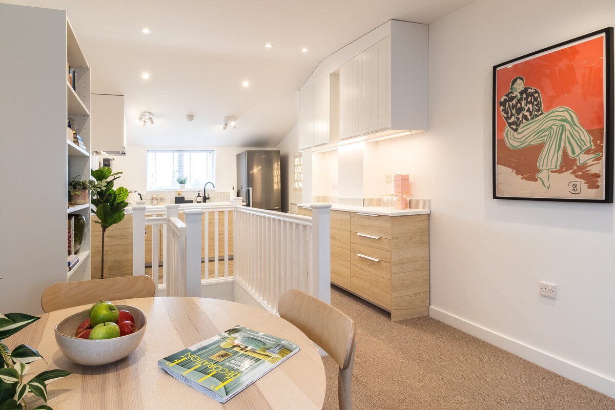 An open-plan kitchen and dining area is presented, showcasing modern cabinetry and a generous countertop. A round dining table is set with a fruit bowl and a magazine. Bright light fills the space from large windows, highlighting the contemporary design and decorative artwork on the wall.